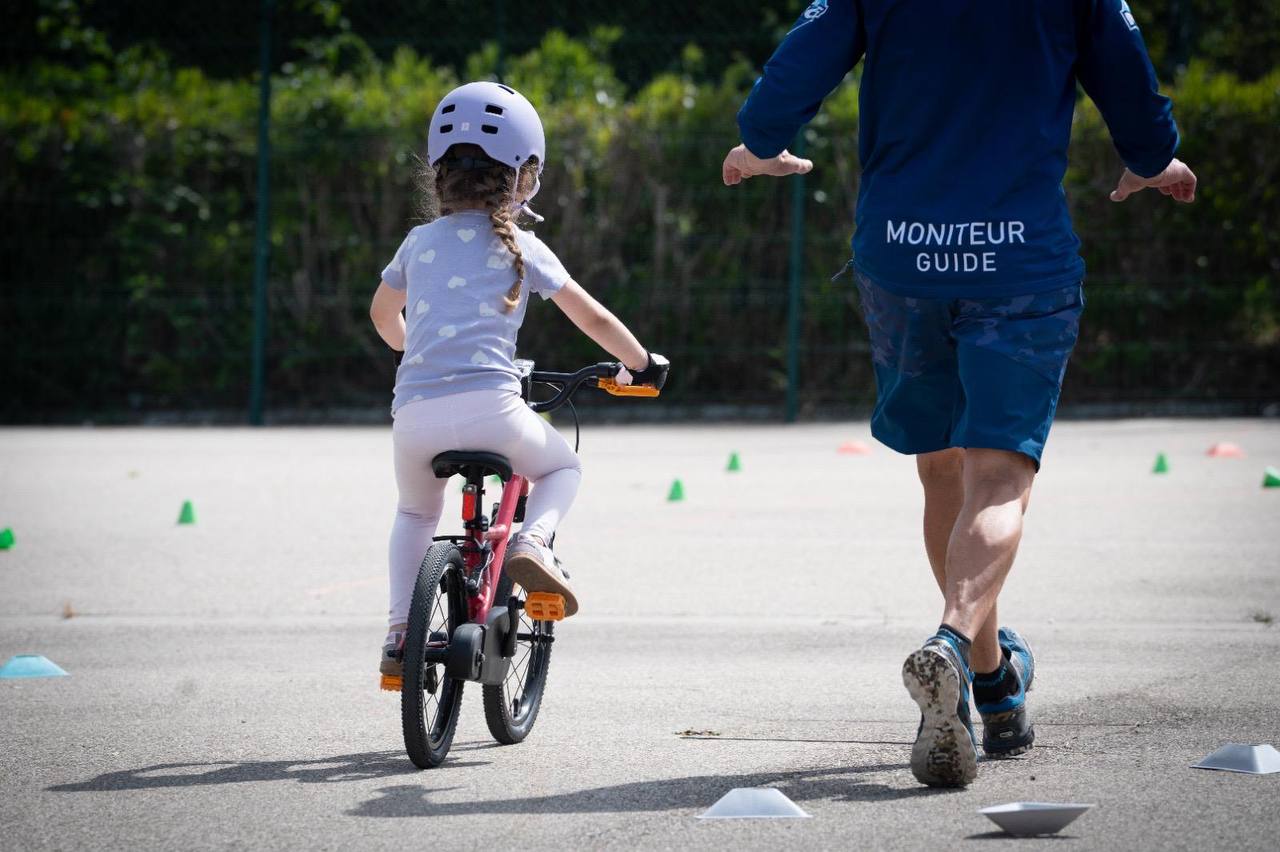 Une fillette casquée apprend à rouler à vélo sans les petites roues, encadrée par un moniteur